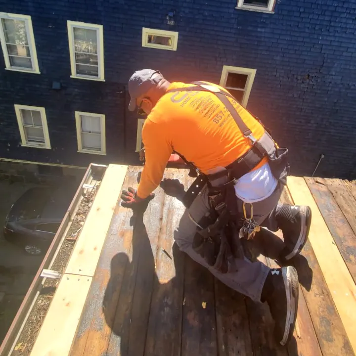 Carpenter in a safety harness laying deck boards on a roof deck between brick buildings