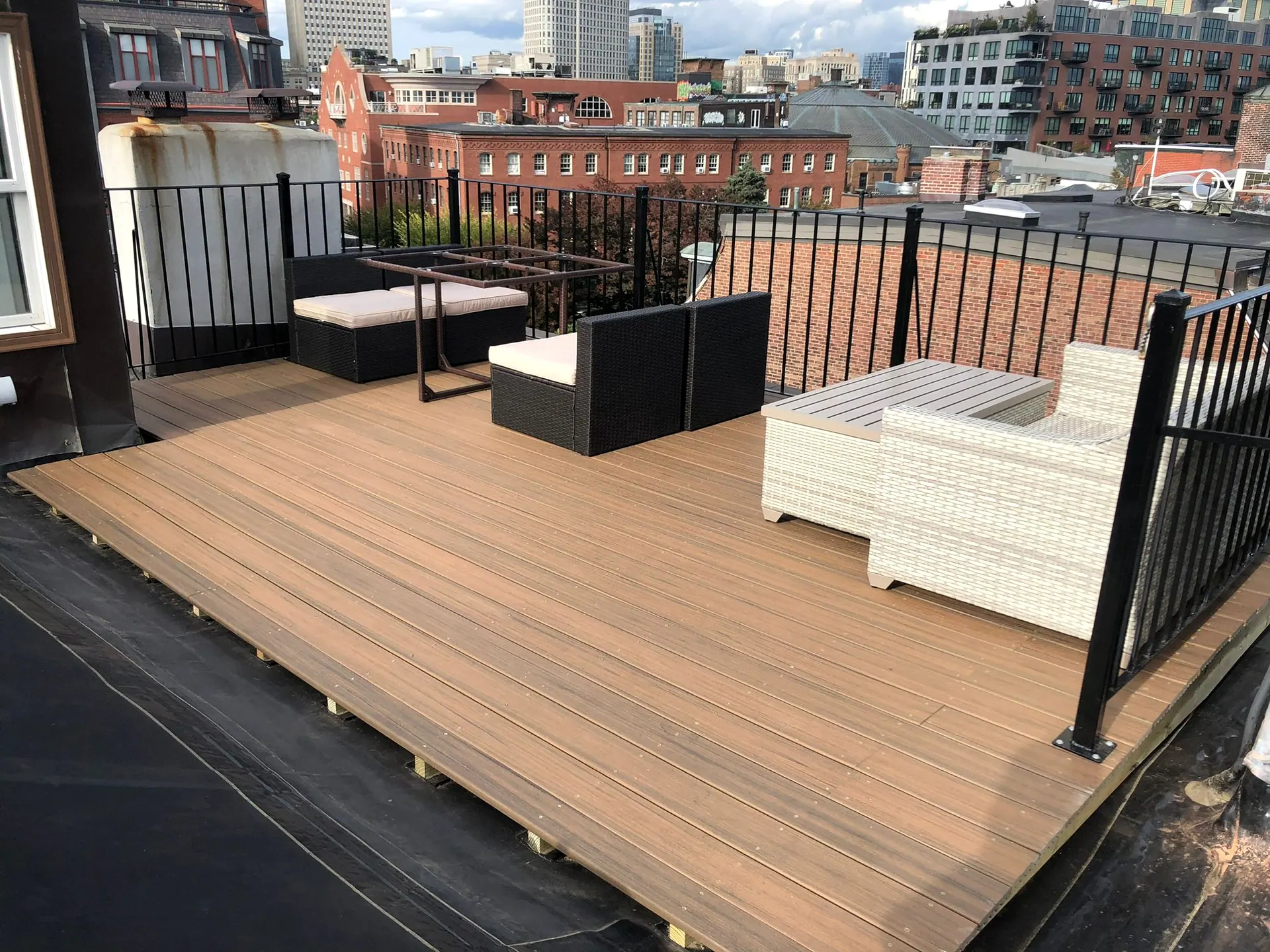 Finished rooftop deck with tan composite boards, black metal railing and outdoor furniture, brick buildings and city towers behind