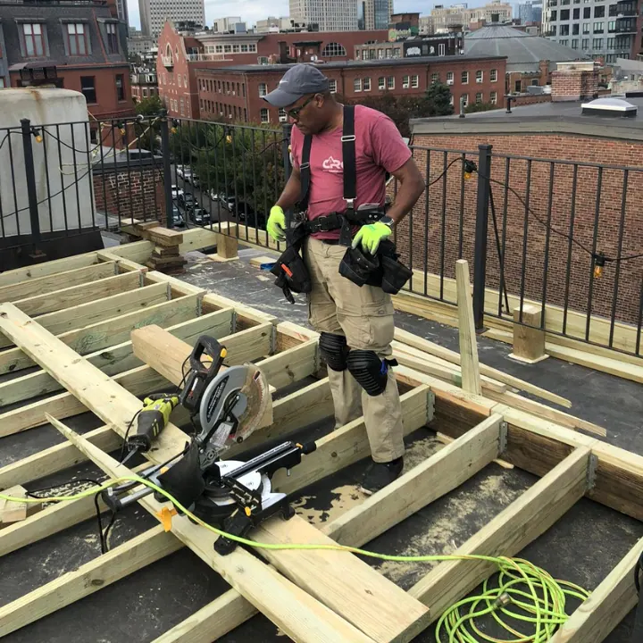 CRC carpenter framing a rooftop deck with pressure-treated joists and a miter saw, city buildings behind